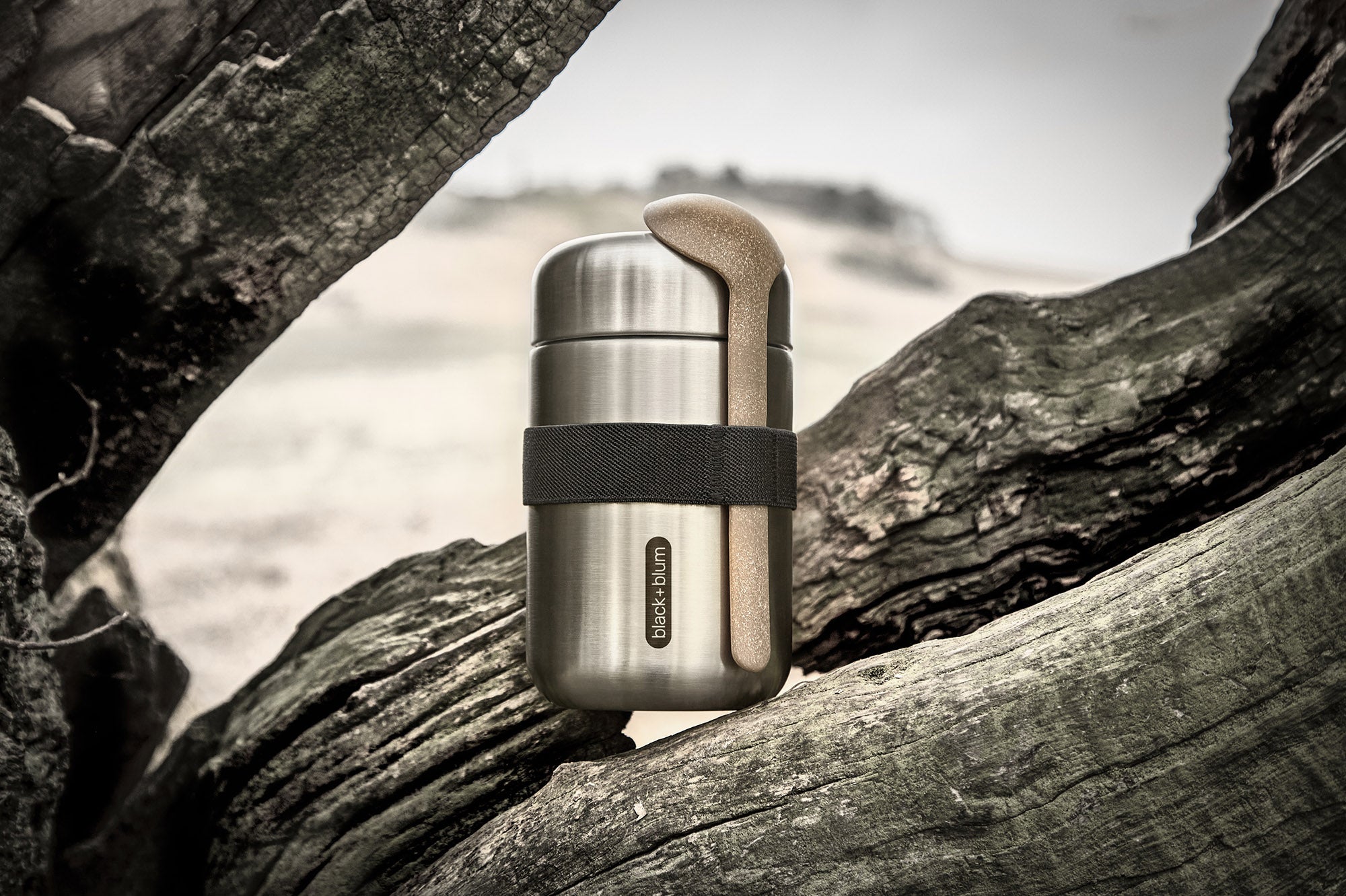 Black + Blum insulated stainless steel food flask with a wooden spoon and black elastic strap resting on a tree