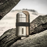 Black + Blum insulated stainless steel food flask with a wooden spoon and black elastic strap resting on a tree