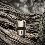 Black + Blum insulated stainless steel food flask with a black elastic strap and wooden spoon resting on a tree