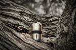 Black + Blum insulated stainless steel food flask with a black elastic strap and wooden spoon resting on a tree