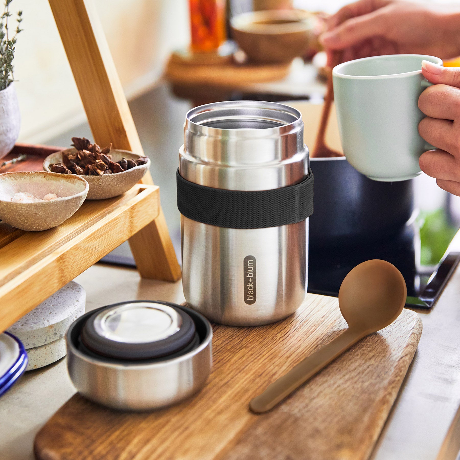 An open Black + Blum insulated stainless steel food flask placed on a small wooden board in a kitchen. An adult's hand can be seen in the background preparing food. The wooden ladle has been placed besides the flask.