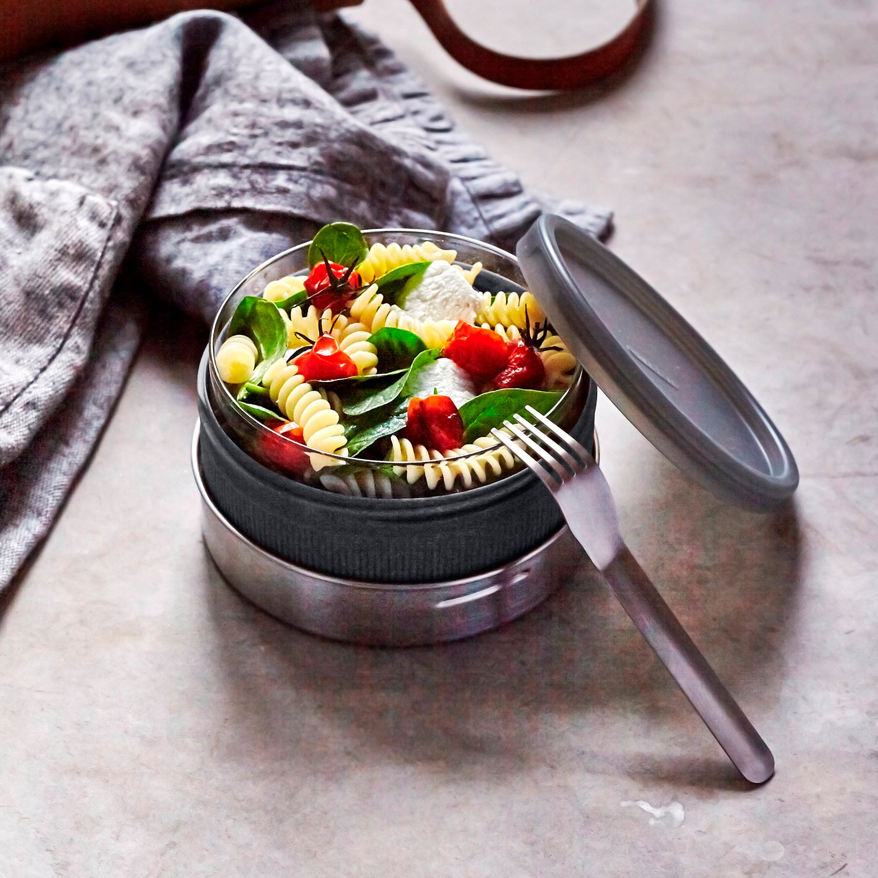 An open Black+Blum Leakproof Microwave Safe Glass Lunch Bowl filled with pasta, basil and tomatoes.  The lid and a stainless steel fork are resting besides it. This lunch box is part of a wide range of reusable stainless steel lunch wares here at Babipur.