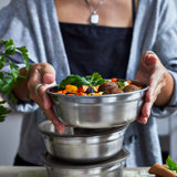 An adult standing over a kitchen worktop with different sized Black + Blum stainless steel bowls stacked on top of eachother