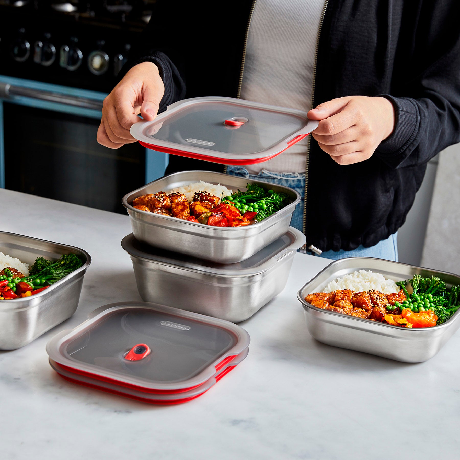 Various sizes of meal prep boxes by Black and Blum in use. The boxes are filled with food and a lid is being placed on one of the boxes.