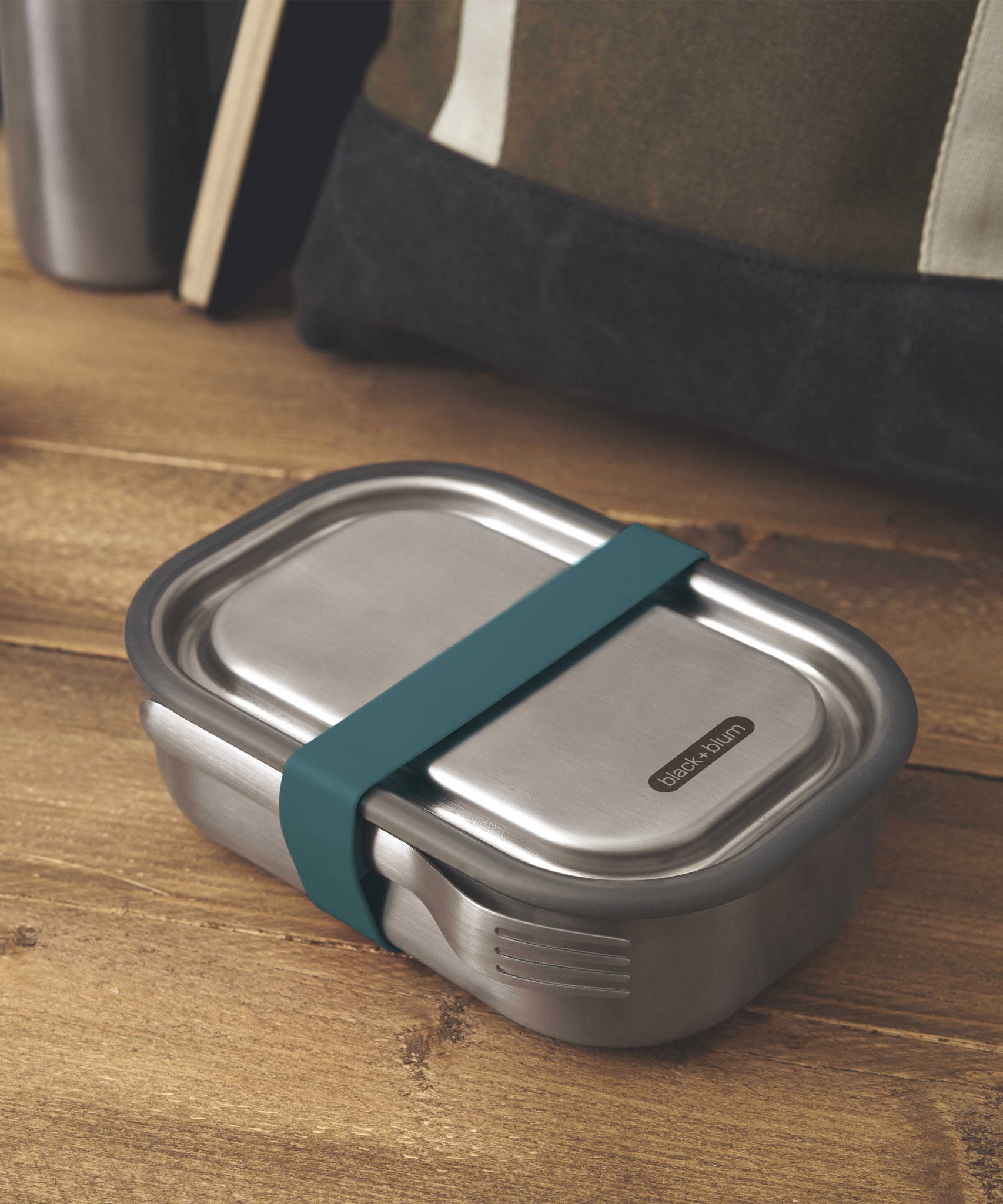 A large Black + Blum stainless steel lunchbox with a ocean blue silicone strap holding a stainless steel fork available at Babipur.