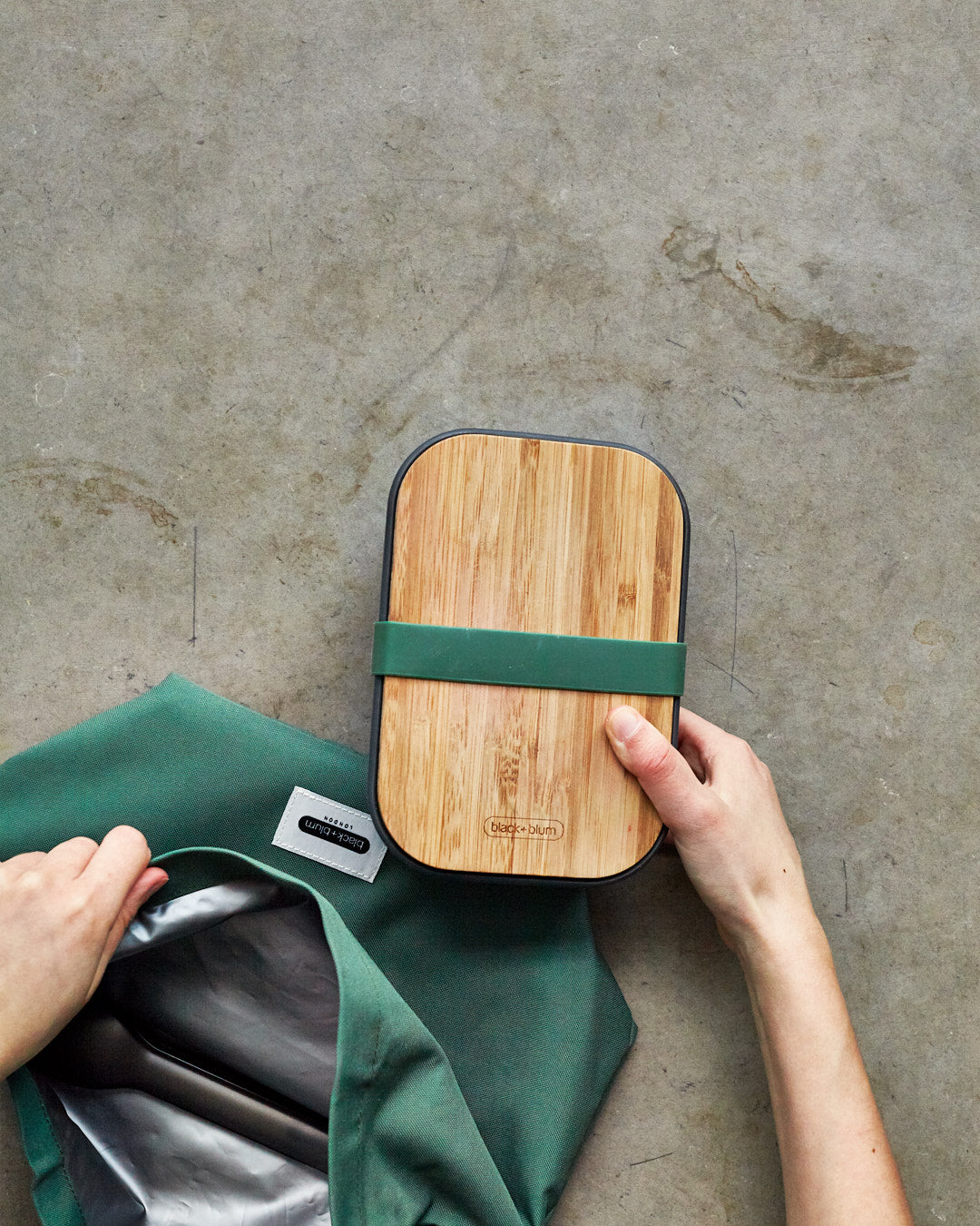 An adult hand holding the Black + Blum stainless steel sandwich box showing the the bamboo lid detail