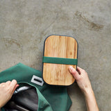 An adult hand holding the Black + Blum stainless steel sandwich box showing the the bamboo lid detail