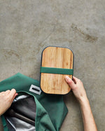 An adult hand holding the Black + Blum stainless steel sandwich box showing the the bamboo lid detail