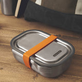 A large Black + Blum stainless steel lunchbox with an  orange silicone strap holding a stainless steel fork available at Babipur.