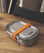 A large Black + Blum stainless steel lunchbox with an  orange silicone strap holding a stainless steel fork available at Babipur.