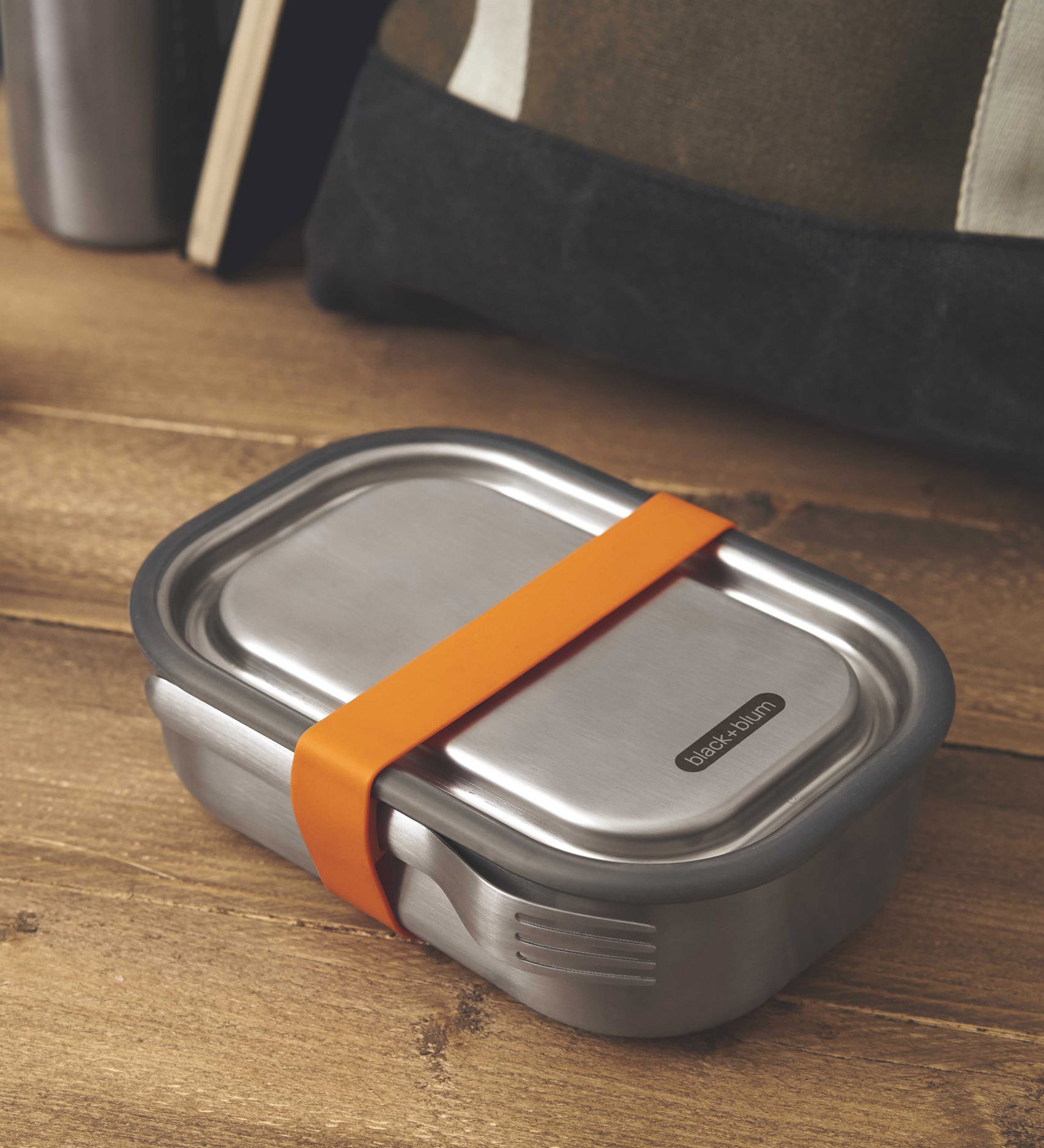 A large Black + Blum stainless steel lunchbox with an  orange silicone strap holding a stainless steel fork available at Babipur.