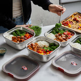 A close up of a adult standing over a kitchen worktop and pouring food into the Black + Blum square stainless steel leakproof microwave safe lunchbox