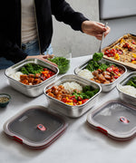 A close up of a adult standing over a kitchen worktop and pouring food into the Black + Blum square stainless steel leakproof microwave safe lunchbox