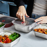 A close up of a adult standing over a kitchen worktop and pressing down on the lid lock on the Black + Blum square stainless steel leakproof microwave safe lunchbox
