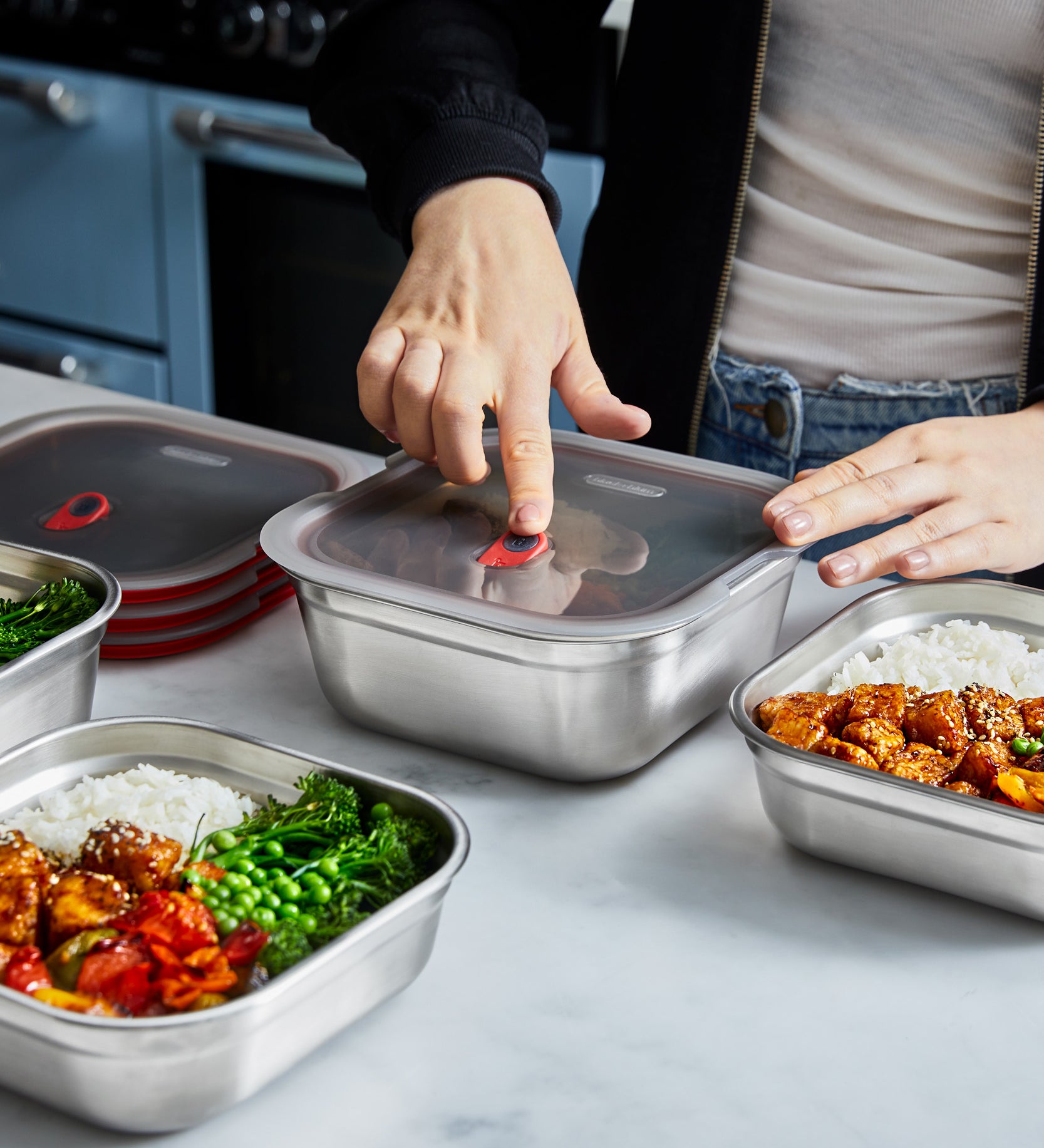 A close up of a adult standing over a kitchen worktop and pressing down on the lid lock on the Black + Blum square stainless steel leakproof microwave safe lunchbox