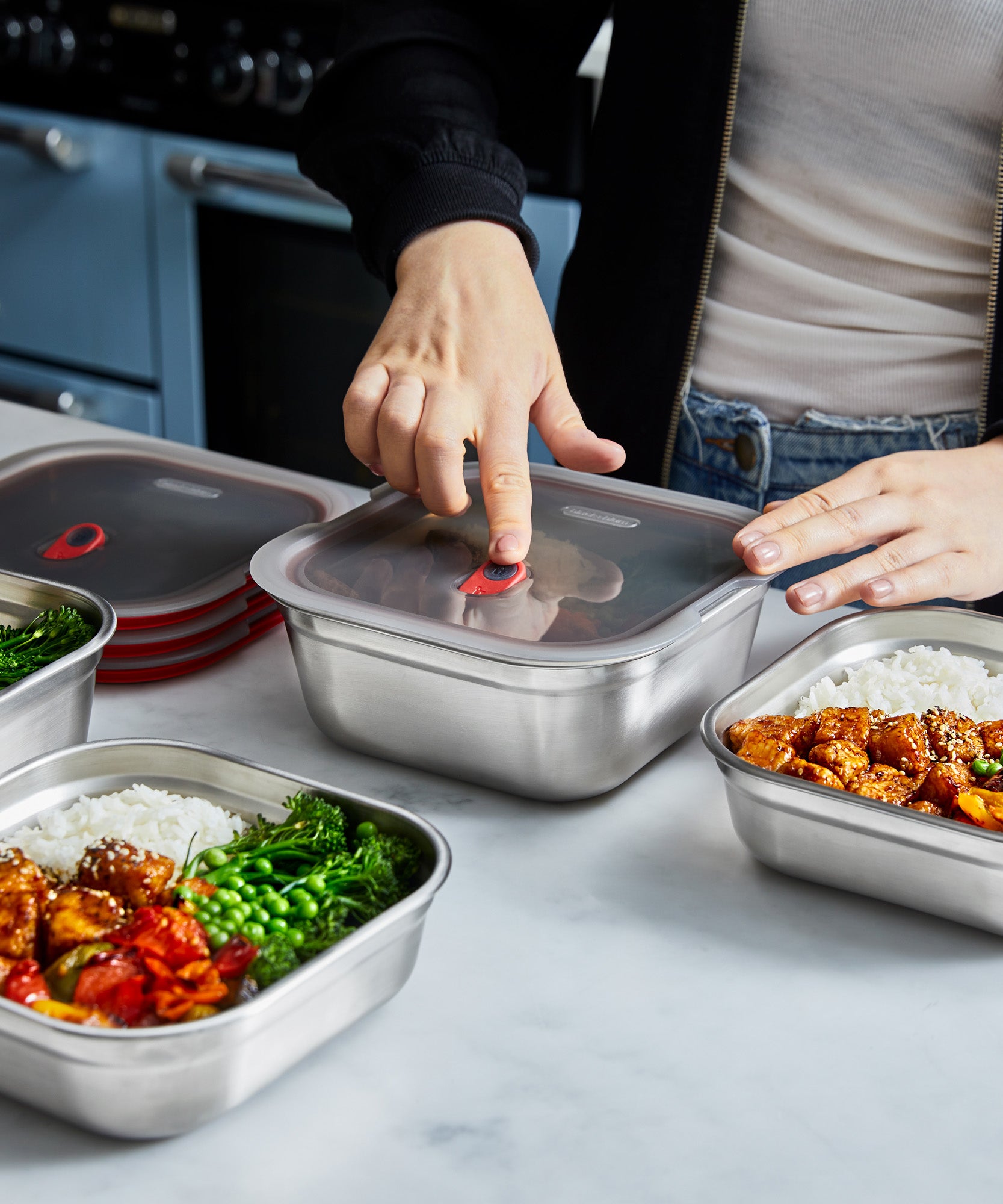 A close up of a adult standing over a kitchen worktop and pressing down on the lid lock on the Black + Blum square stainless steel leakproof microwave safe lunchbox