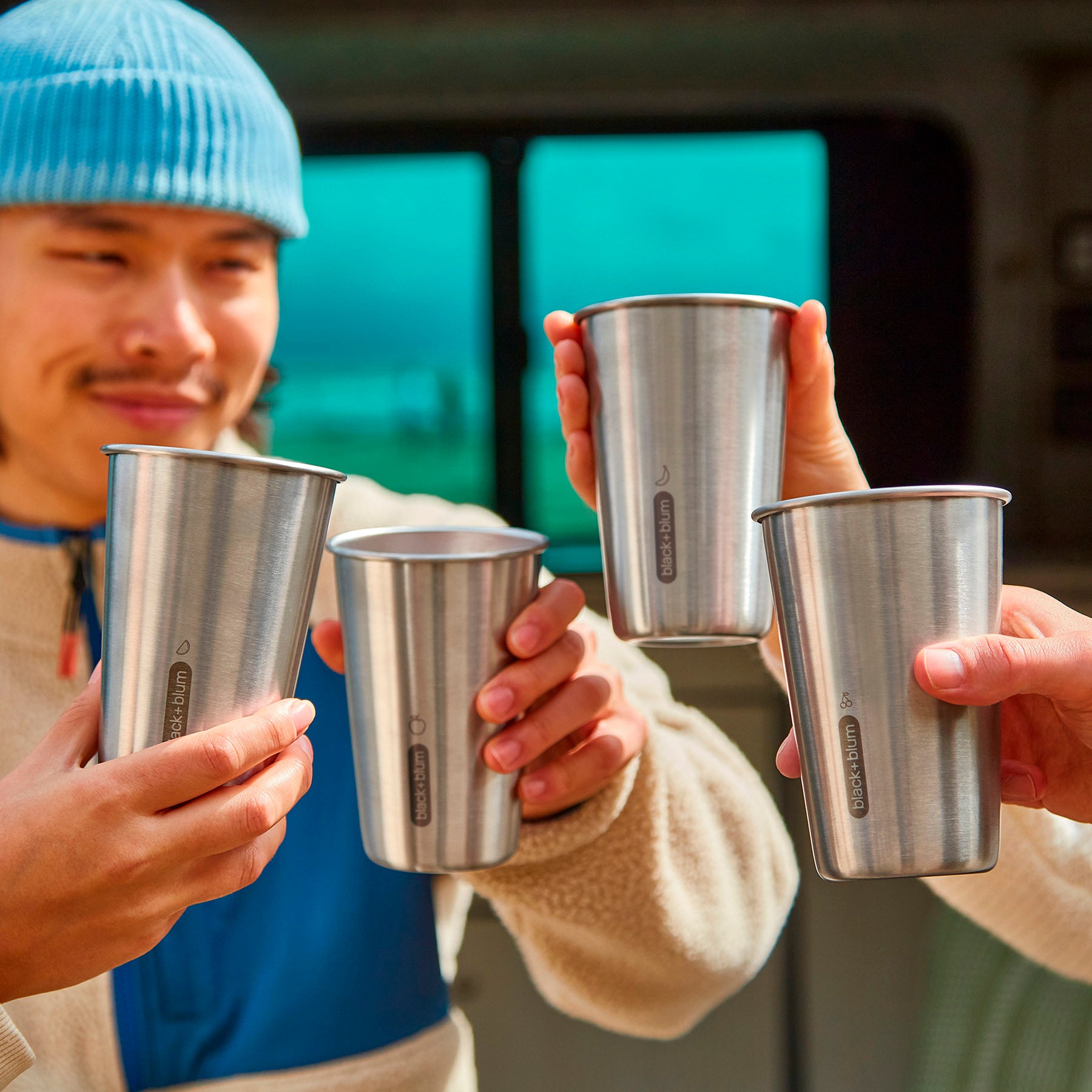 A close up of 4 stainless steel Black and Blum cups in use. A close up of people's hand's holding the cups in a cheers motion.