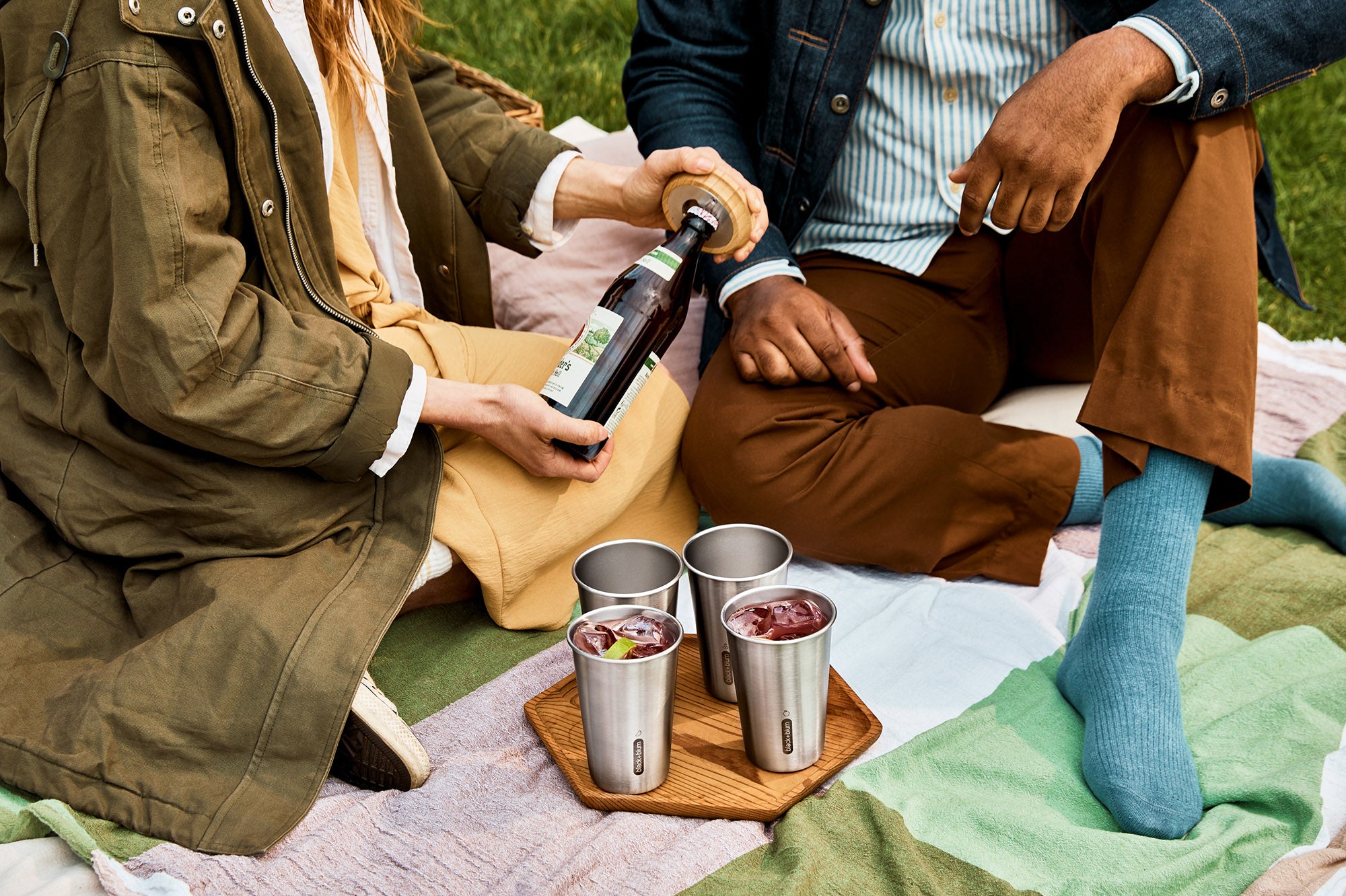 A close up of a couple of adults having a drink using the  Black + Blum stainless steel cups and showing the bamboo lid bottle opener