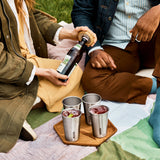 A close up of a couple of adults having a drink using the  Black + Blum stainless steel cups and showing the bamboo lid bottle opener