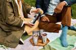 A close up of a couple of adults having a drink using the  Black + Blum stainless steel cups and showing the bamboo lid bottle opener