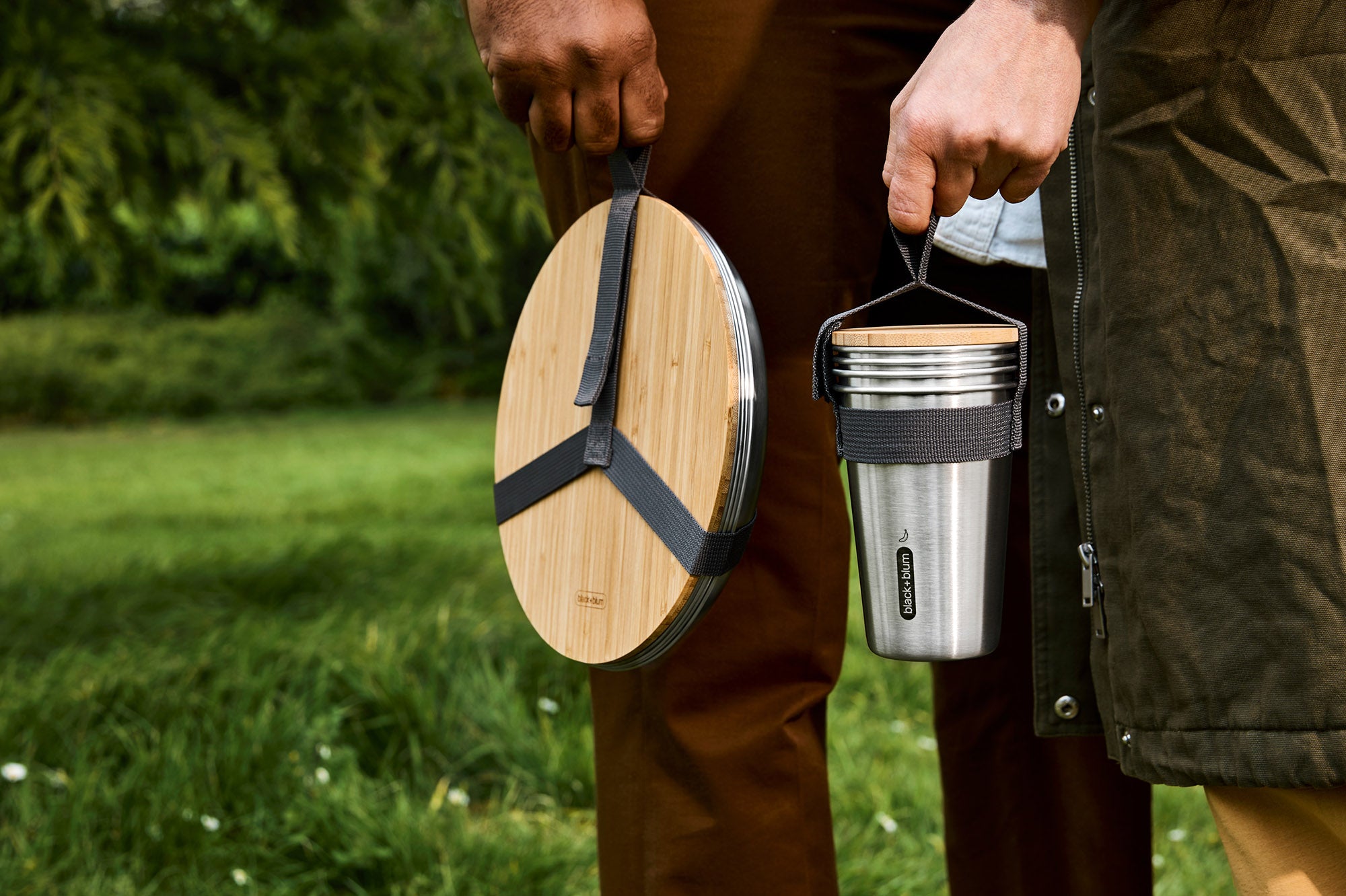 A close up of an adult holding the Black + Blum stainless steel plate and cup set by the handy black strap handle