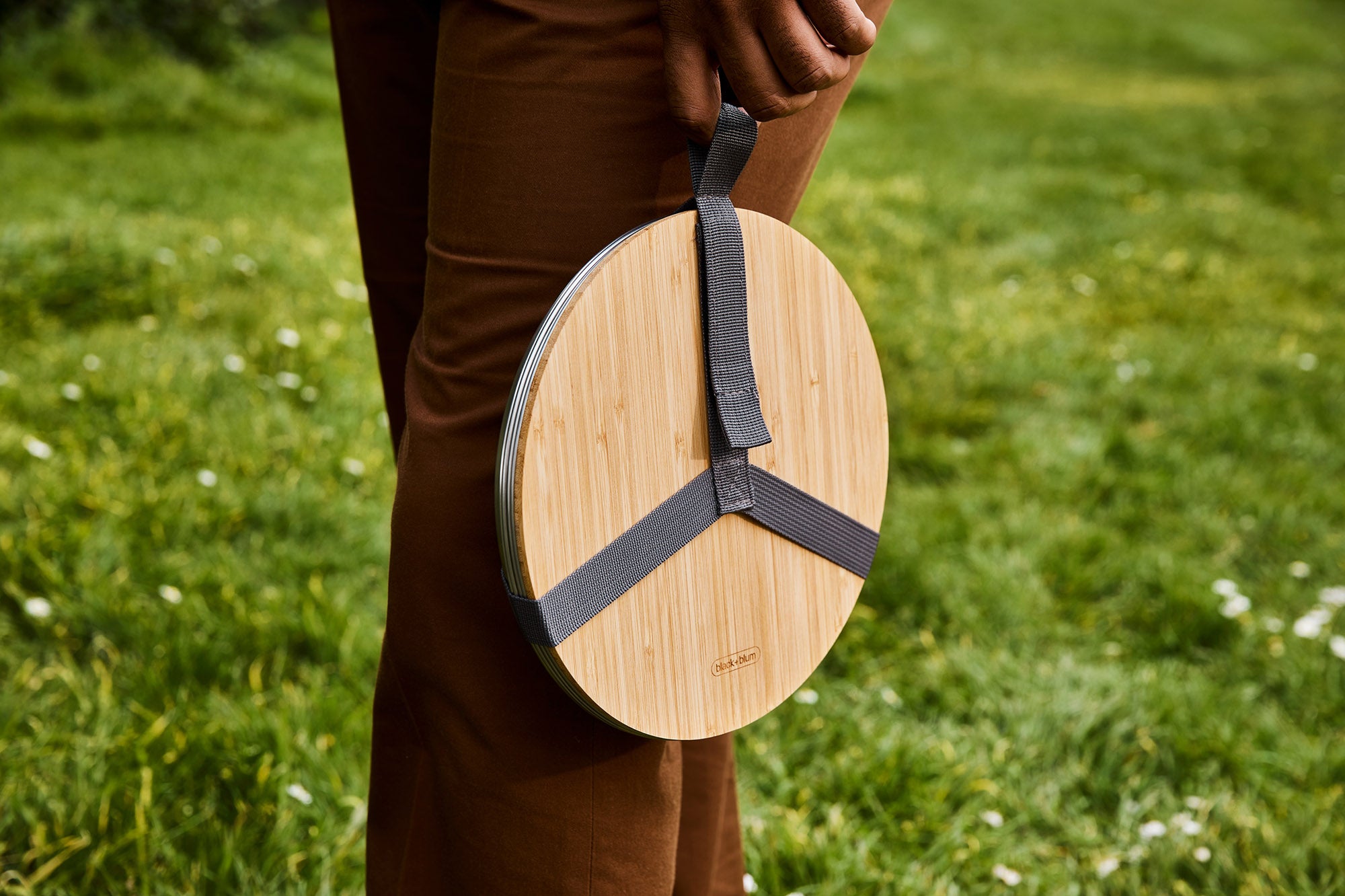 A close up of an adult holding the Black + Blum stainless steel plate by the handy black strap handle and showing the protective bamboo lid detail