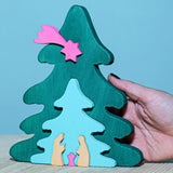 A Bumbu Christmas Tree Nativity Puzzle in a Fuchsia colour way placed on a wooden surface. An adult's hand can be seen on the side for scale.