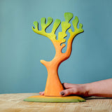 Adults hand holding the Bumbu Big Dino Tree placed on a wooden surface in front of a blue wall
