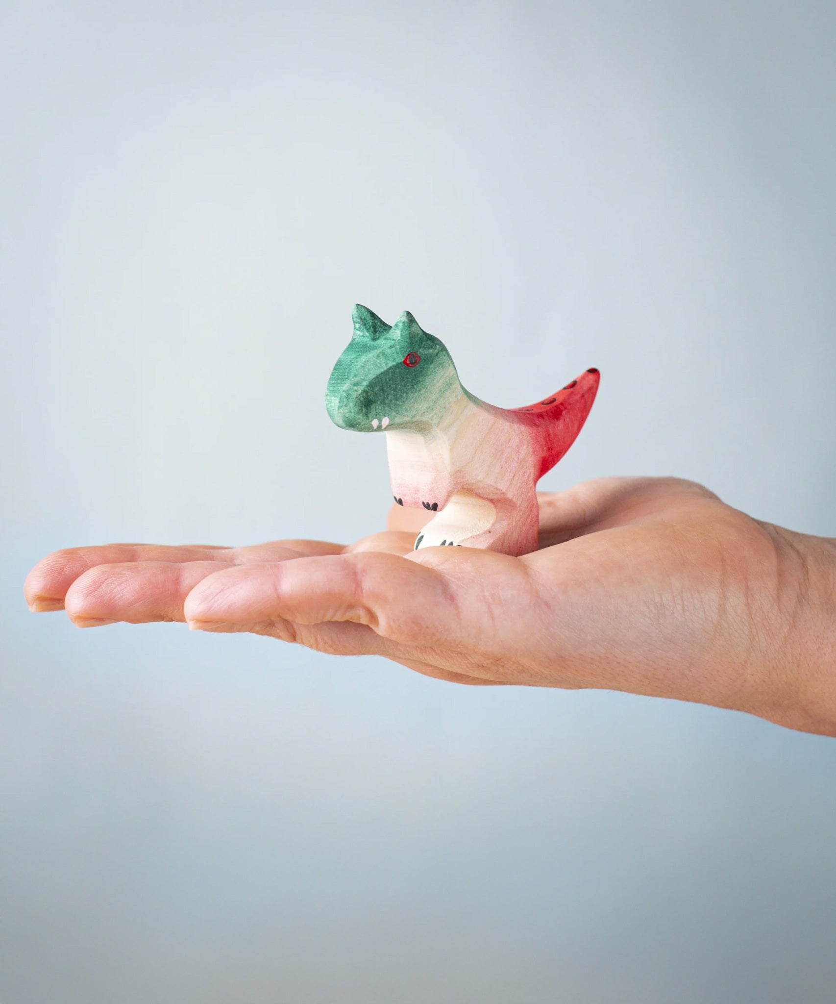 A Bumbu Toys handmade wooden Baby Carnotaurus dinosaur figure shown on an adult's hand.