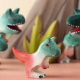 A Bumbu Toys Baby Carnotaurus dinosaur placed at the front of a small world play scene with other handmade wooden toys from Bumbu in the background.