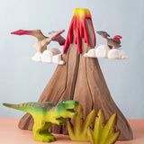 A small world play scene featuring handmade wooden toys from a brand called Bumbu. There are Pterandons and a t-rex standing on and around a wooden volcano and green coloured wooden foliage.