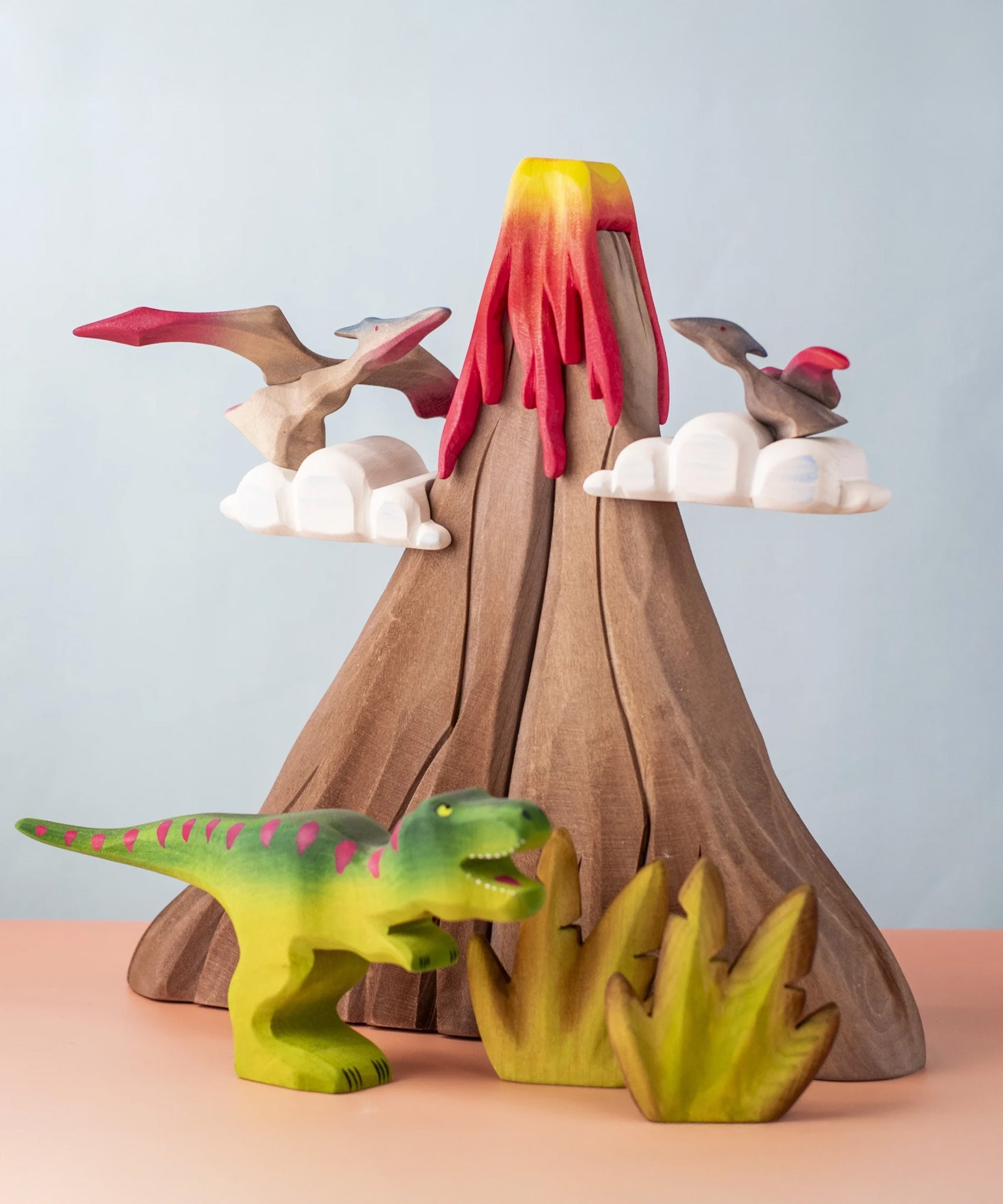 A small world play scene featuring handmade wooden toys from a brand called Bumbu. There are Pterandons and a t-rex standing on and around a wooden volcano and green coloured wooden foliage.