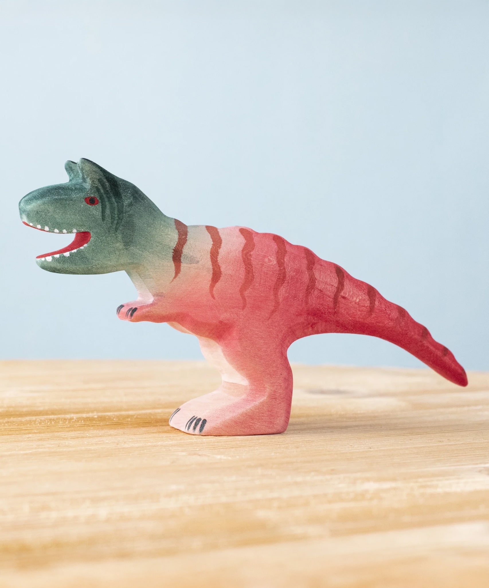 Bumbu Toys Big Carnotaurus dinosaur wooden figure placed on a wooden surface. The handmade dinosaur figure is part of a wide range of wooden animal figures here at Babipur.