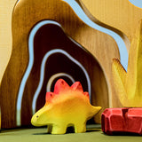 A wooden Bumbu baby stegosaurus stood in front of wooden cave arches and a red coloured wooden rock.