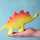 A large wooden Bumbu stegosaurus figure balancing on an adult's hand against a blue background.