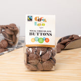 Bag of Cocoa Loco organic milk chocolate buttons on a wooden table in front of a glass cup and metal scoop filled with chocolate buttons