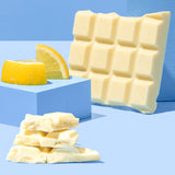 The Divine White Chocolate Lemon Cheesecake Dessert Bar shown unwrapped and broken up with a pile of the pieces shown at the front with pieces of lemon behind.