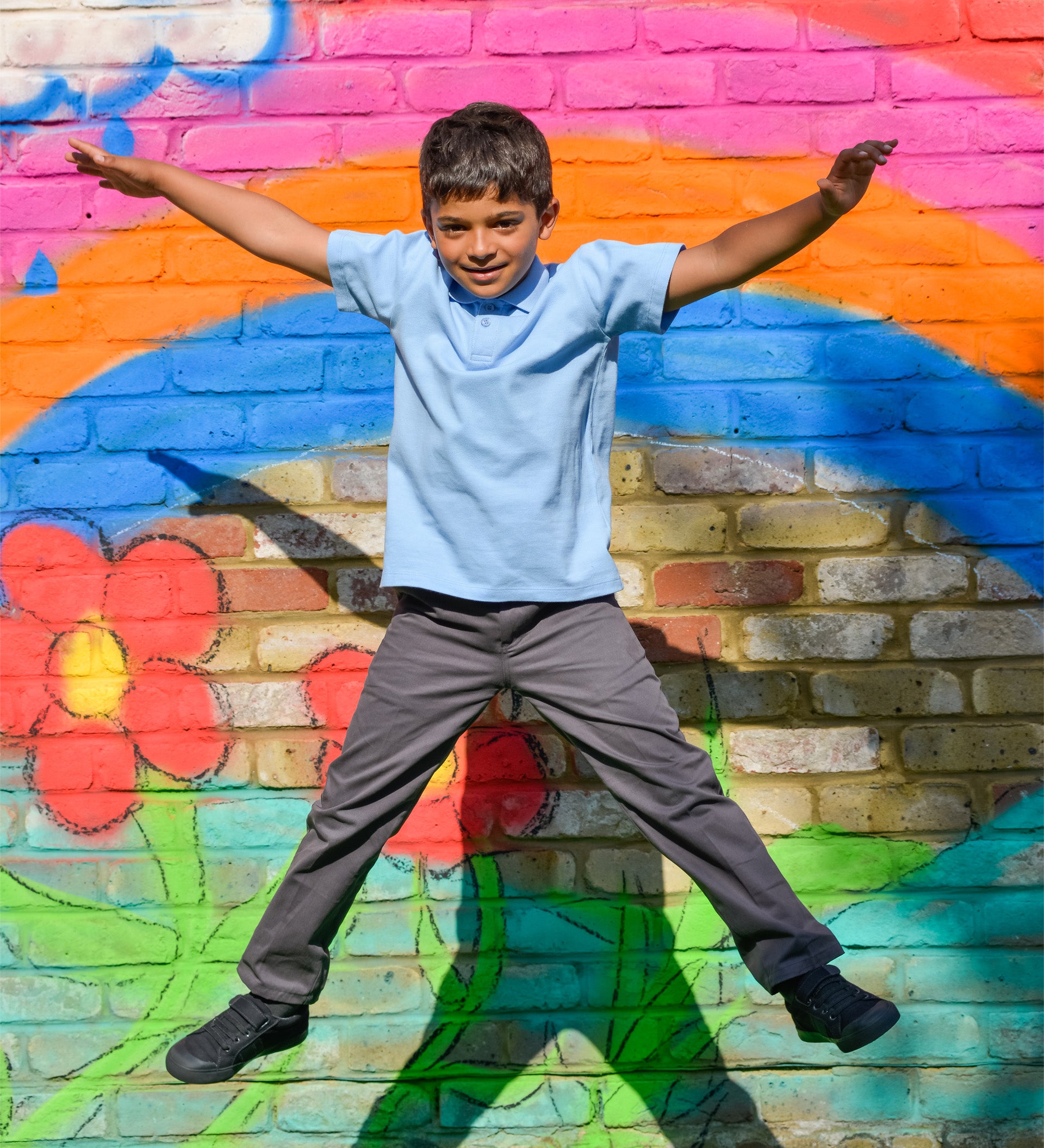 A school boy making a star jump in the air, wearing the Eco Outfitters organic and fair trade cotton polo shirt and school trousers.