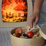 A child's hand taking a white coloured mushroom from the box of Erzi forest mushrooms. There is a lit Toverlux Magic lamp in the background with Autumn gnome toadstool picnic artwork by artist Vogel Geluk inside.