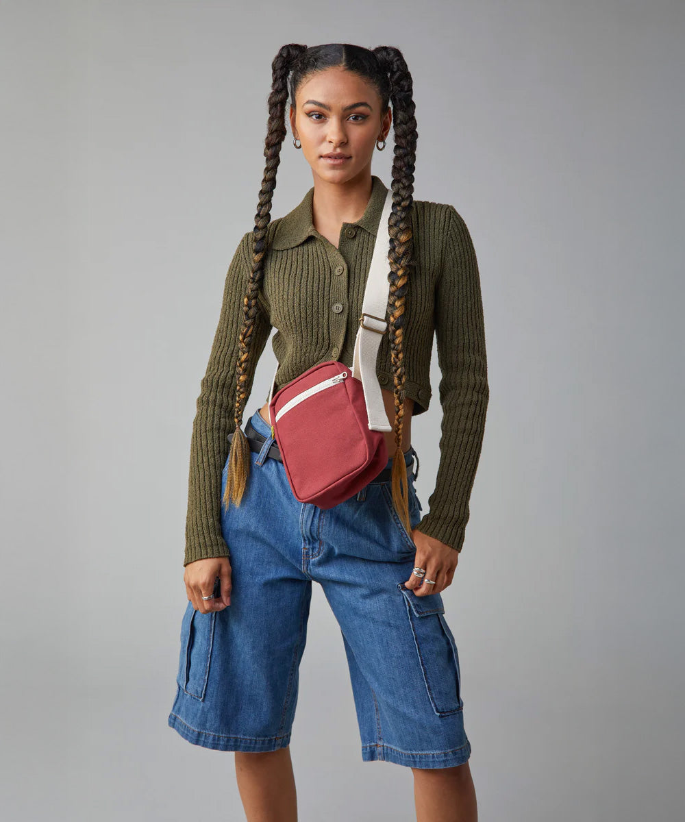 A woman with braided hair, wearing the burgundy organic cotton Mini Crossbody bag from Fluf, a sporty and sleek bag for snacks, travel, and festivals