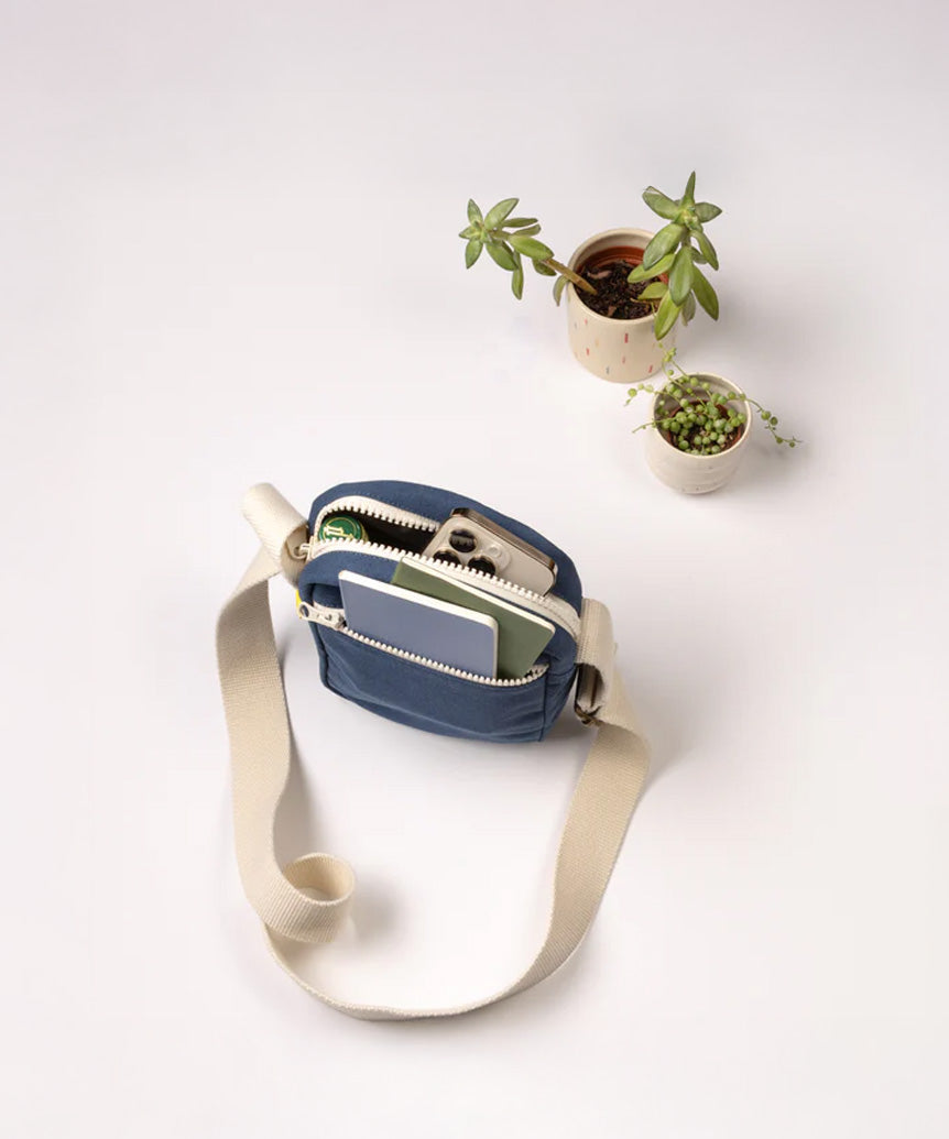 A top view of the Fluf organic cotton mini crossbody bag in navy, holding an iPhone and small notebook in the separated lined pockets.