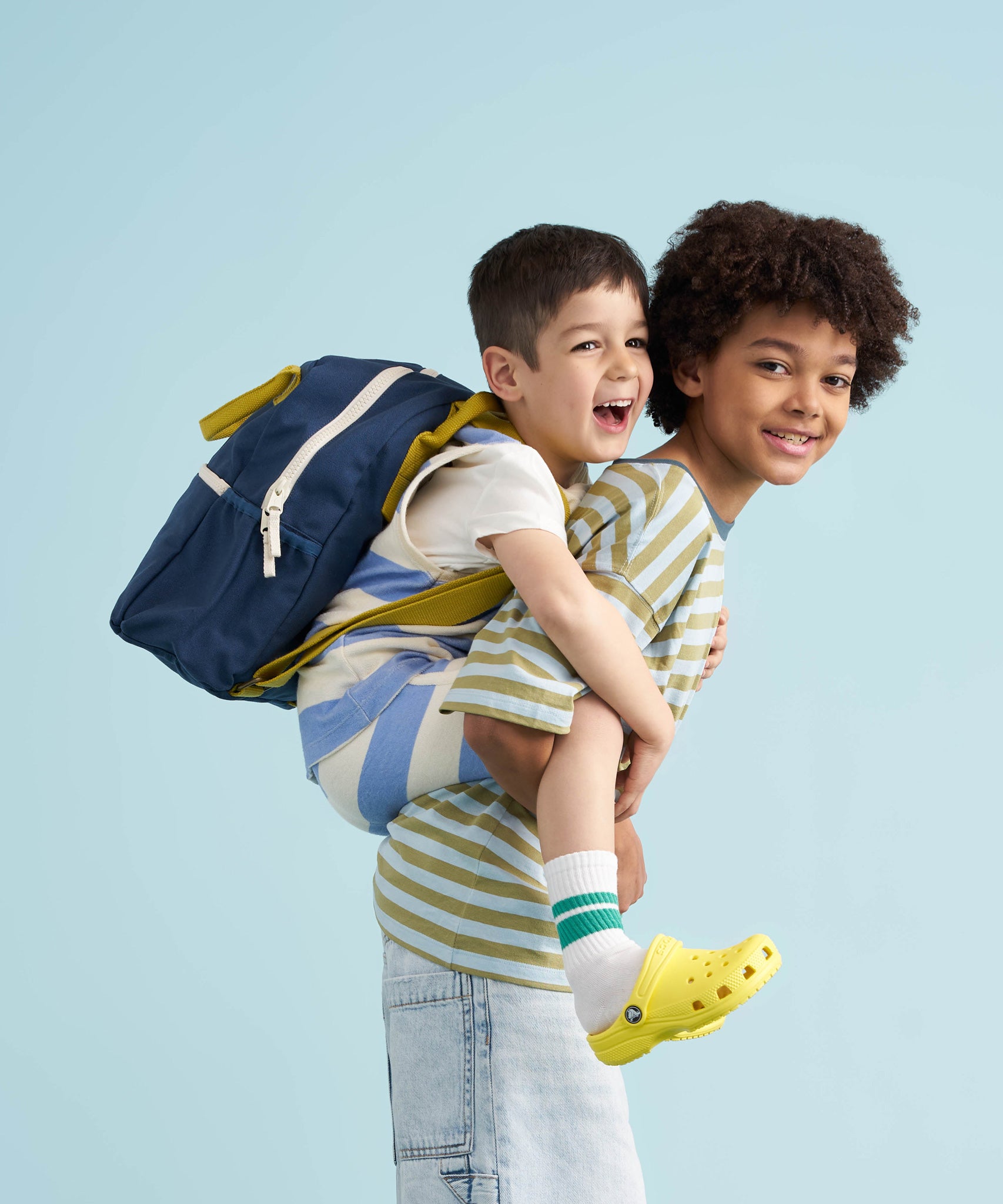 A child wearing a navy coloured Fluf junior backpack whilst having a piggy back from an older child. Both are smiling.