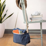 The Fluf organic cotton crossbody lunch bag in navy blue with a white strap, hanging on the back of a chair and filled with books.