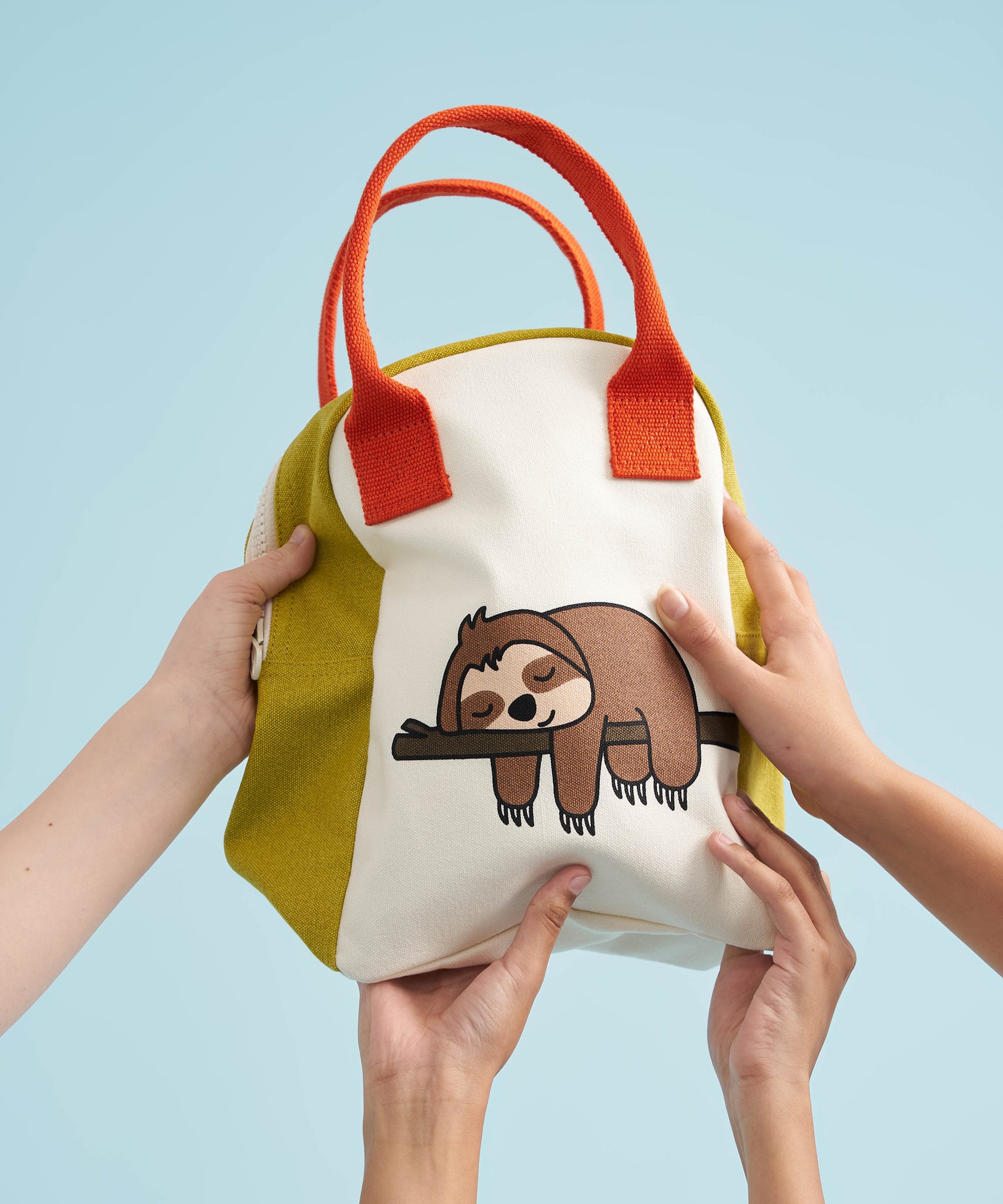 The Fluf organic cotton zippered lunch bag, featuring a sloth design on the front, orange handles and green side panels. Hands can be seen holding up the bag.