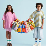 Two children holding up 9 different design of the Fluf organic cotton zippered lunch bags on their arms. They are stood holding hands and the bags are all lined up and hung on their arms by the handles.