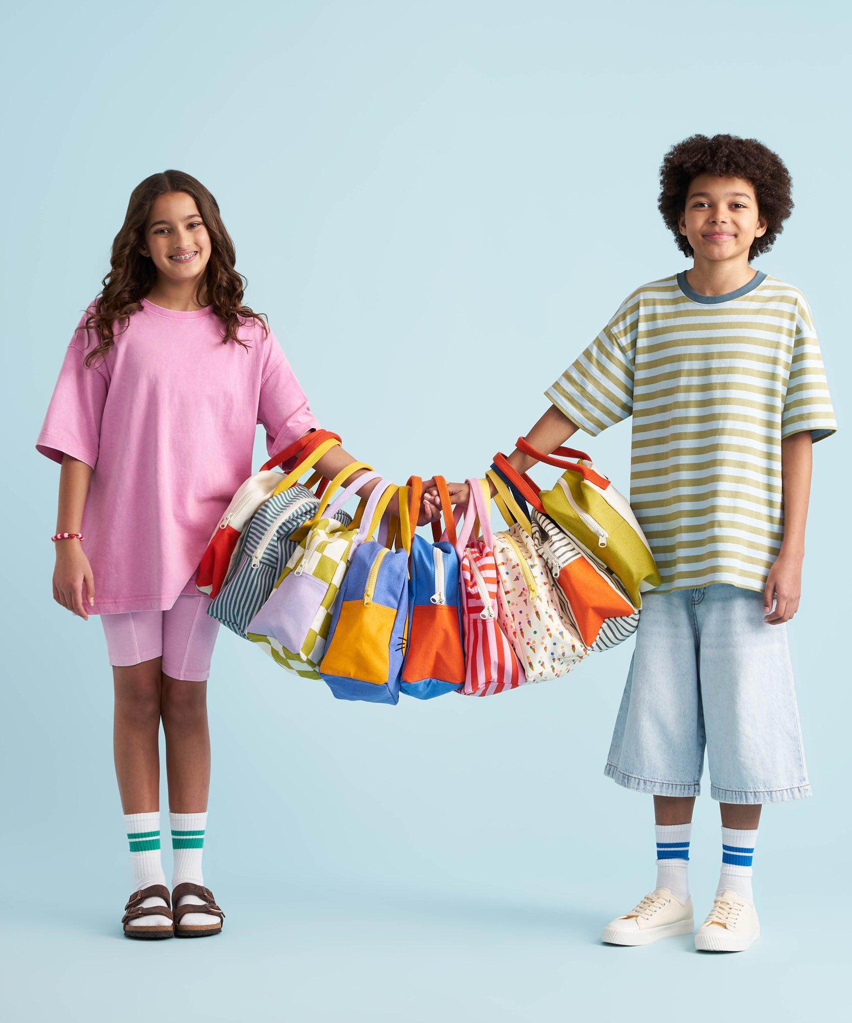 Two children holding up 9 different design of the Fluf organic cotton zippered lunch bags on their arms. They are stood holding hands and the bags are all lined up and hung on their arms by the handles.