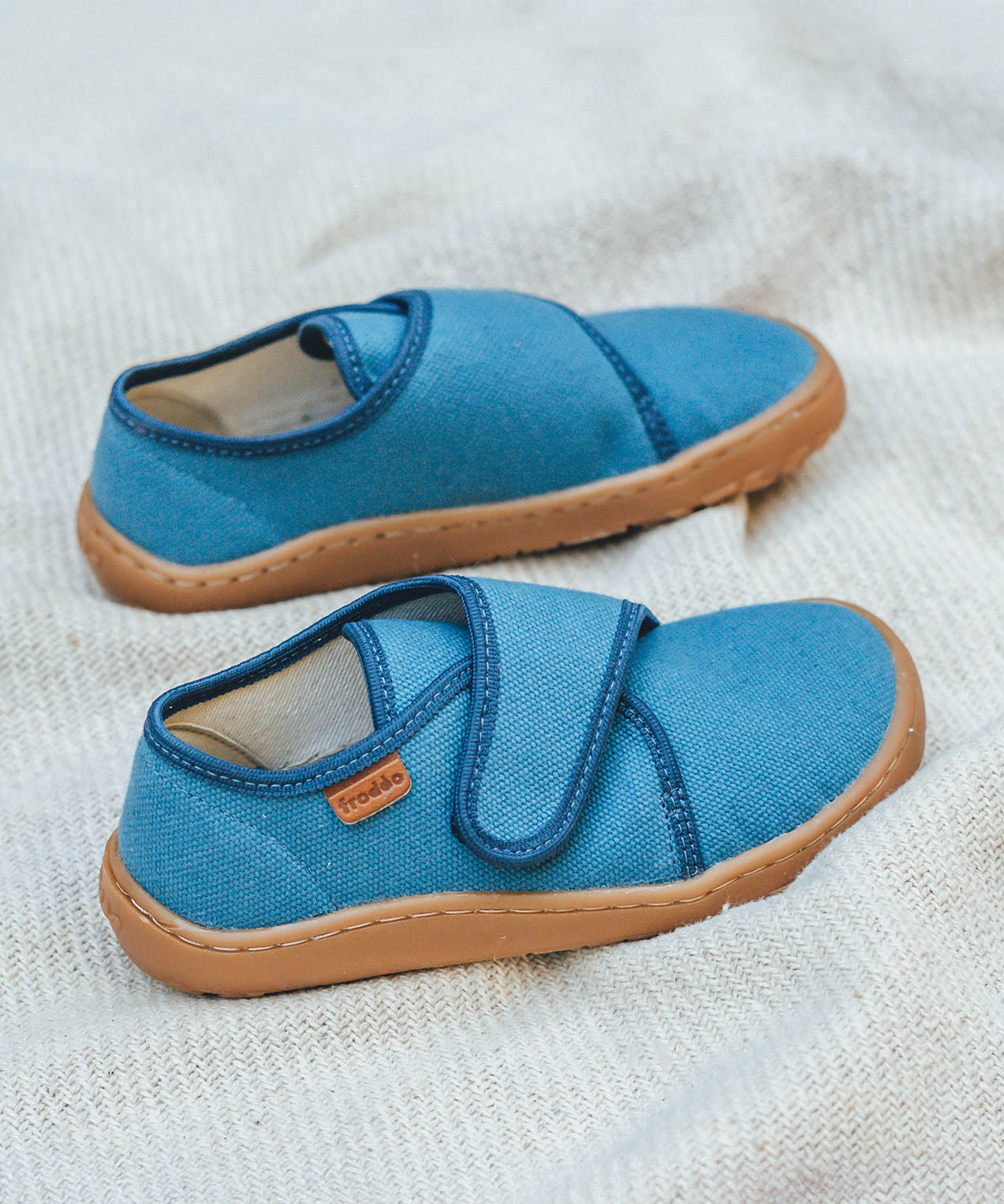 Froddo barefoot kids slipper in denim blue available at Babipur standing on a soft pale blanket. A canvas style slipper shoe in a light neutral colour with a velcro fastener for a secure fit and a dark brown grippy sole