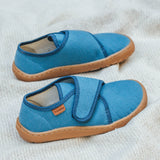 Froddo barefoot kids slipper in denim blue available at Babipur standing on a soft pale blanket. A canvas style slipper shoe in a light neutral colour with a velcro fastener for a secure fit and a dark brown grippy sole