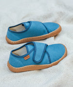 Froddo barefoot kids slipper in denim blue available at Babipur standing on a soft pale blanket. A canvas style slipper shoe in a light neutral colour with a velcro fastener for a secure fit and a dark brown grippy sole