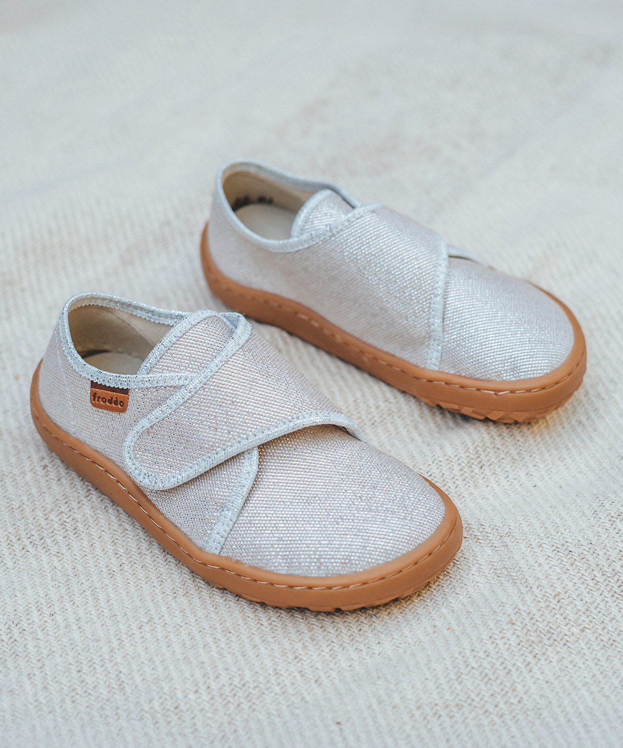 Froddo barefoot kids slipper in gold shine available at Babipur standing on a soft pale blanket. A canvas style slipper shoe in a light neutral colour with a velcro fastener for a secure fit and a dark brown grippy sole